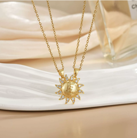 Duo de colliers soleil dorés en acier inoxydable, pendentifs ronds sertis de pierres scintillantes, présentés sur assiette blanche.